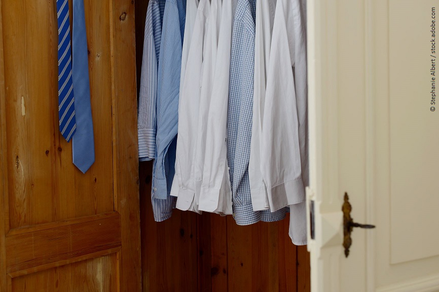 Verschiedene Herrenhemden hängen in einem Holzschrank. In der Tür hängen zwei Krawatten. 